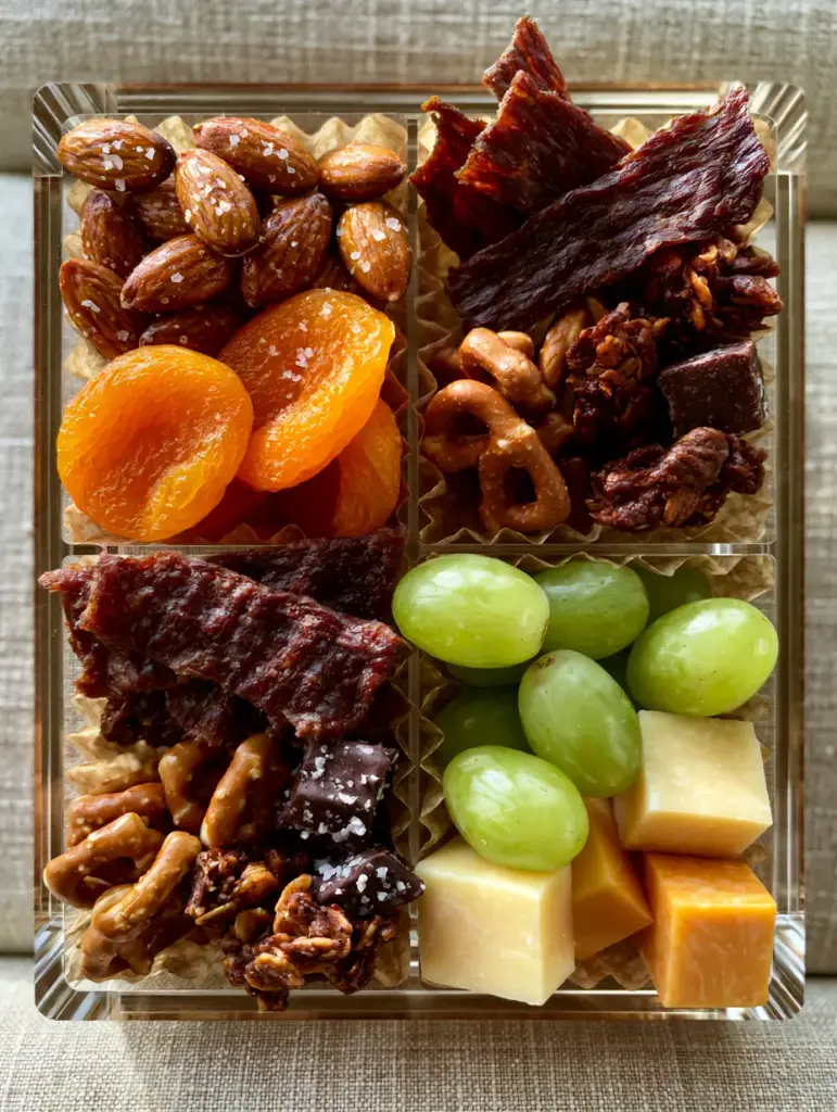 Healthy Road Trip Snacks: The Ultimate High-Protein Travel Box Guide 2 Top-down lifestyle shot of a clear multi-compartment organizer filled with healthy road trip snacks on a linen car seat.
