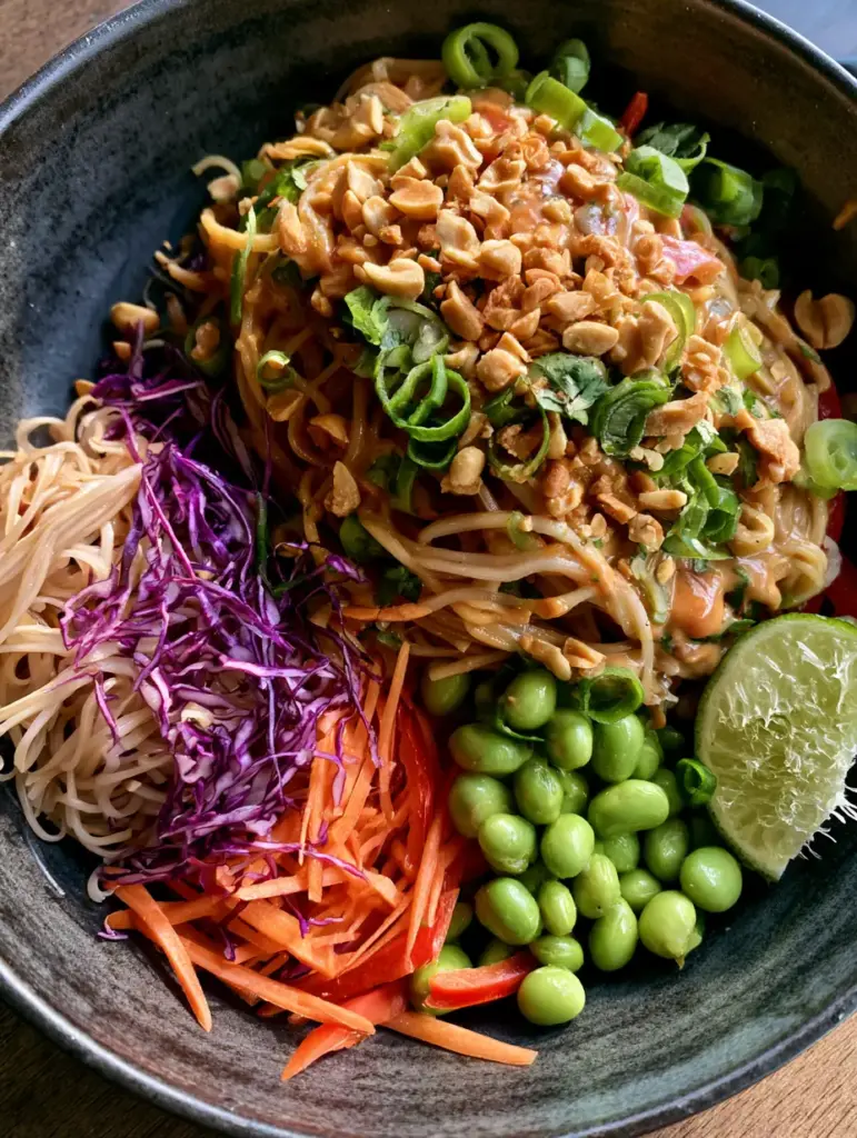 Peanut Pasta Salad with Velvety Ginger Dressing 1 Close up of glossy tan peanut sauce coating rice noodles with crushed peanuts and scallions in a dark charcoal bowl.