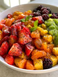 Close-up 45-degree angle of honey lime fruit salad showing the thick, reflective translucent glaze and poppy seeds on kiwi and blackberries.