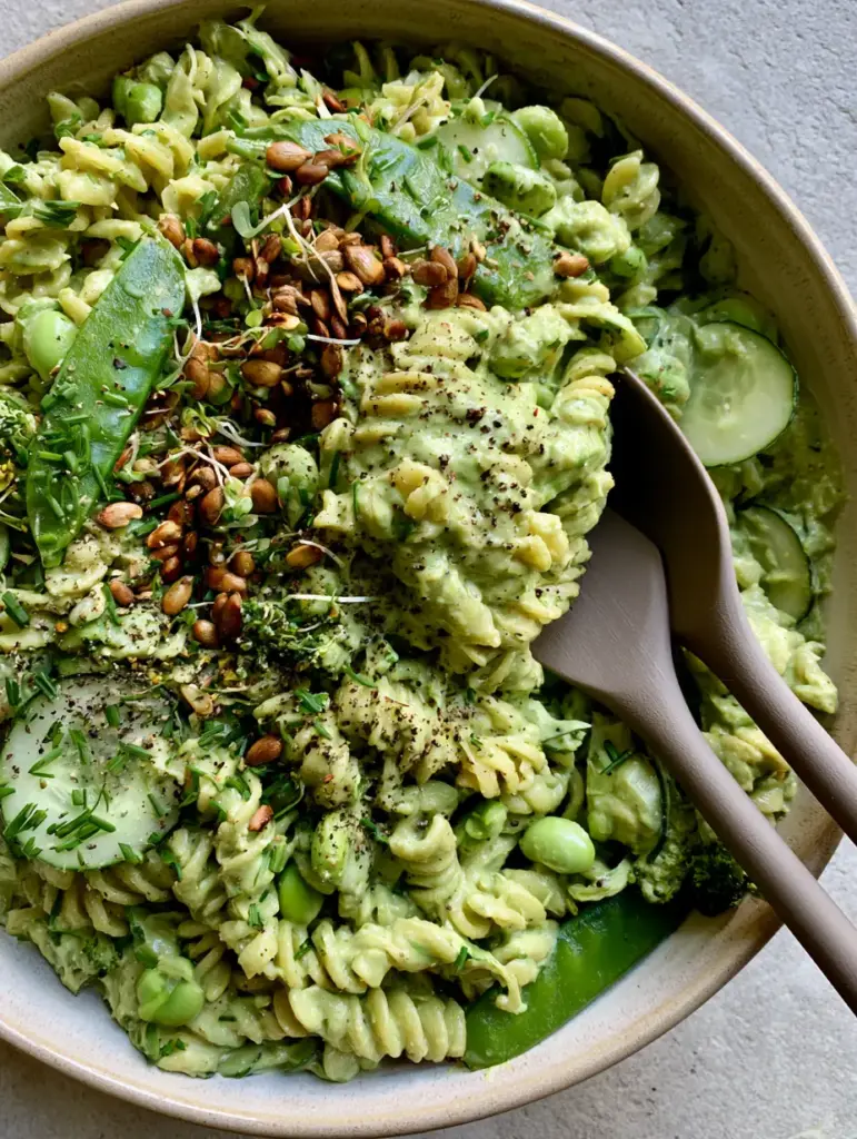 Green Goddess Pasta Salad with Creamy Avocado and Fresh Herbs 1 Overhead view of Green Goddess Pasta Salad garnished with pumpkin seeds, microgreens, and sliced cucumbers on a stone countertop.